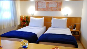 Habitación de hotel con dos camas, cabecero de madera, mesita de noche con un teléfono y un objeto decorativo, y una mesa con un adorno de cristal y un plato.
