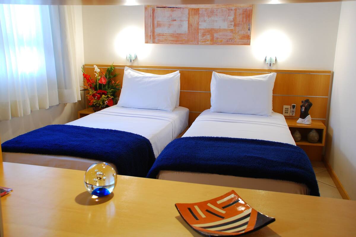 Une chambre d’hôtel avec deux lits, une tête de lit en bois, une table de chevet avec un téléphone et un objet décoratif, et une table sur laquelle se trouvent un ornement en verre et une assiette.