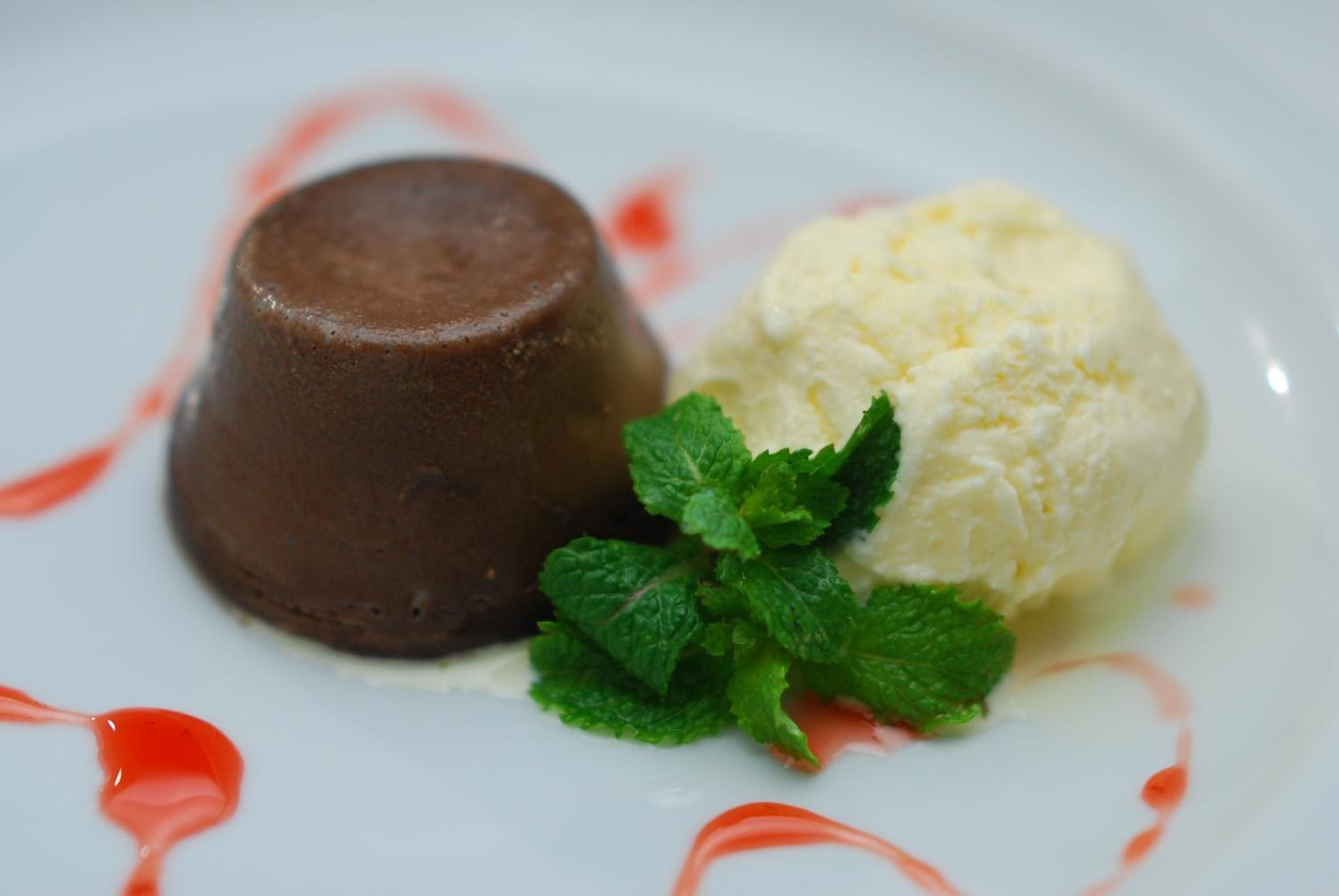 A dessert consisting of a chocolate pudding and a scoop of vanilla ice cream, garnished with mint leaves and drizzled with red syrup.