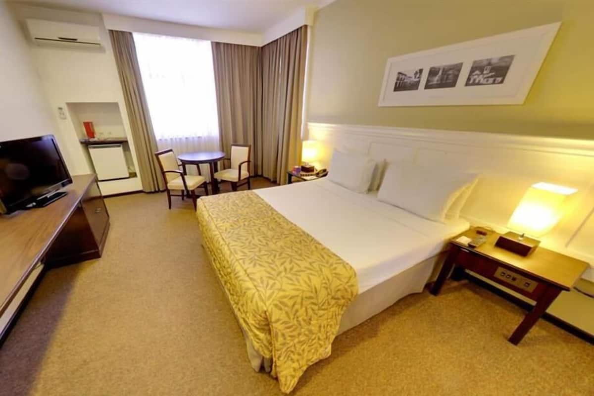 Ein Hotelzimmer mit Bett, Nachttischen, Schreibtisch, Fernseher und einem Fenster mit Vorhängen.
