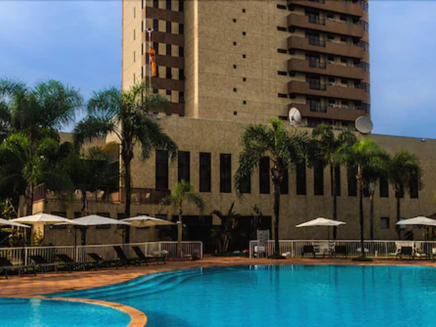 A swimming pool with lounge chairs and umbrellas, surrounded by palm trees and a tall building with balconies.