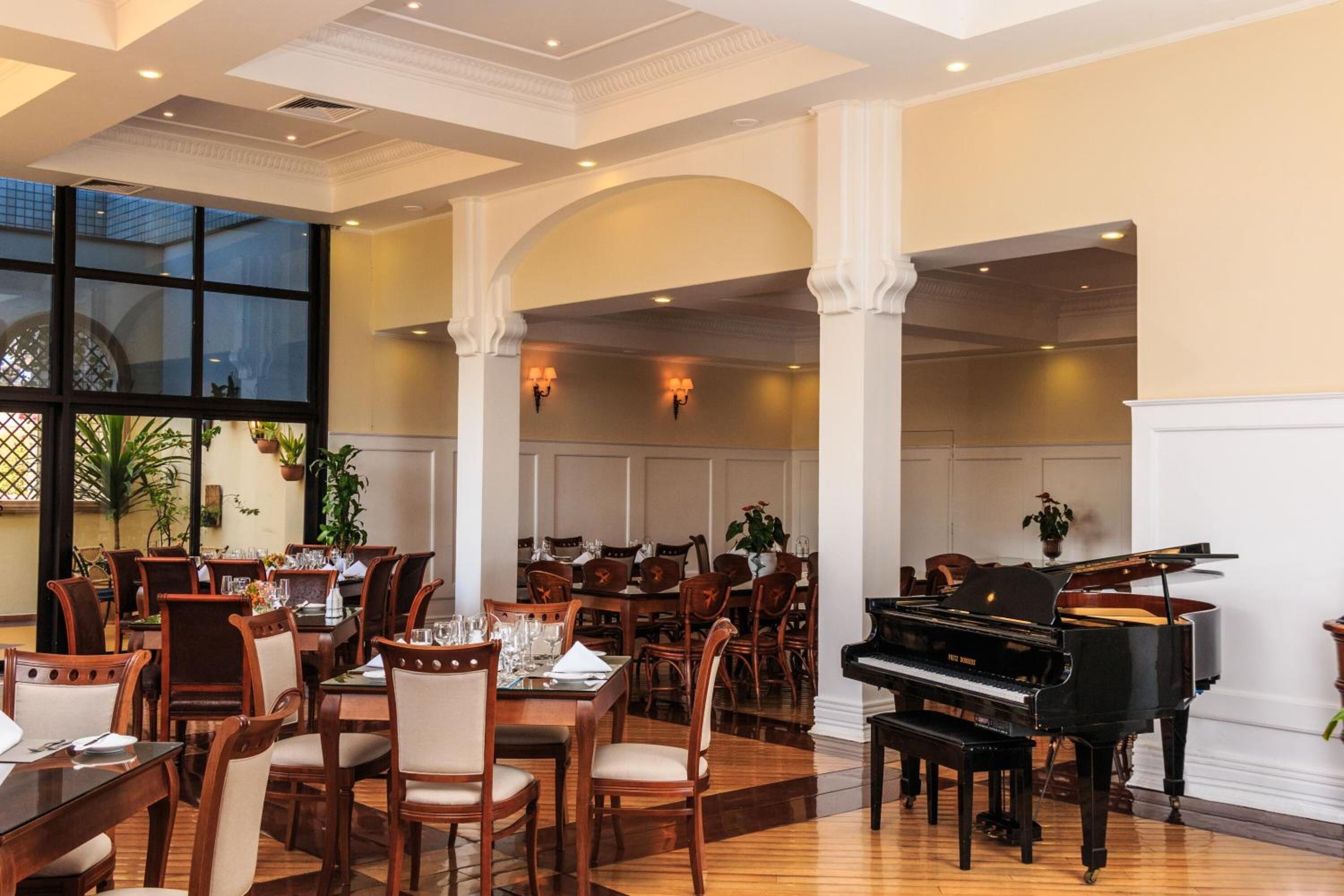 Uma ampla sala de jantar com várias mesas e cadeiras, um piano de cauda e grandes janelas com vista para o exterior.