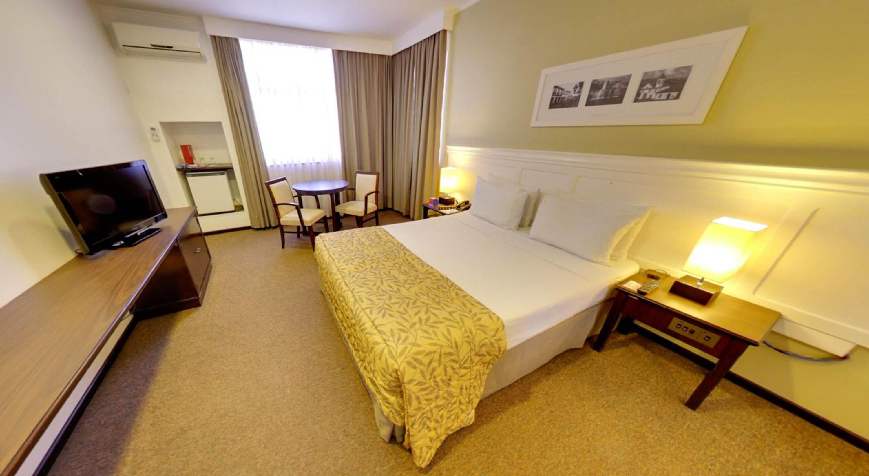 Quarto de hotel com cama, mesa de cabeceira, luminária, televisão, escrivaninha, cadeira e janela com cortinas.