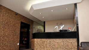 A reception area with a modern design, featuring a reception desk with a black top and a patterned lower section. There is a large mirror above the desk, and a brown leather chair is visible to the right.