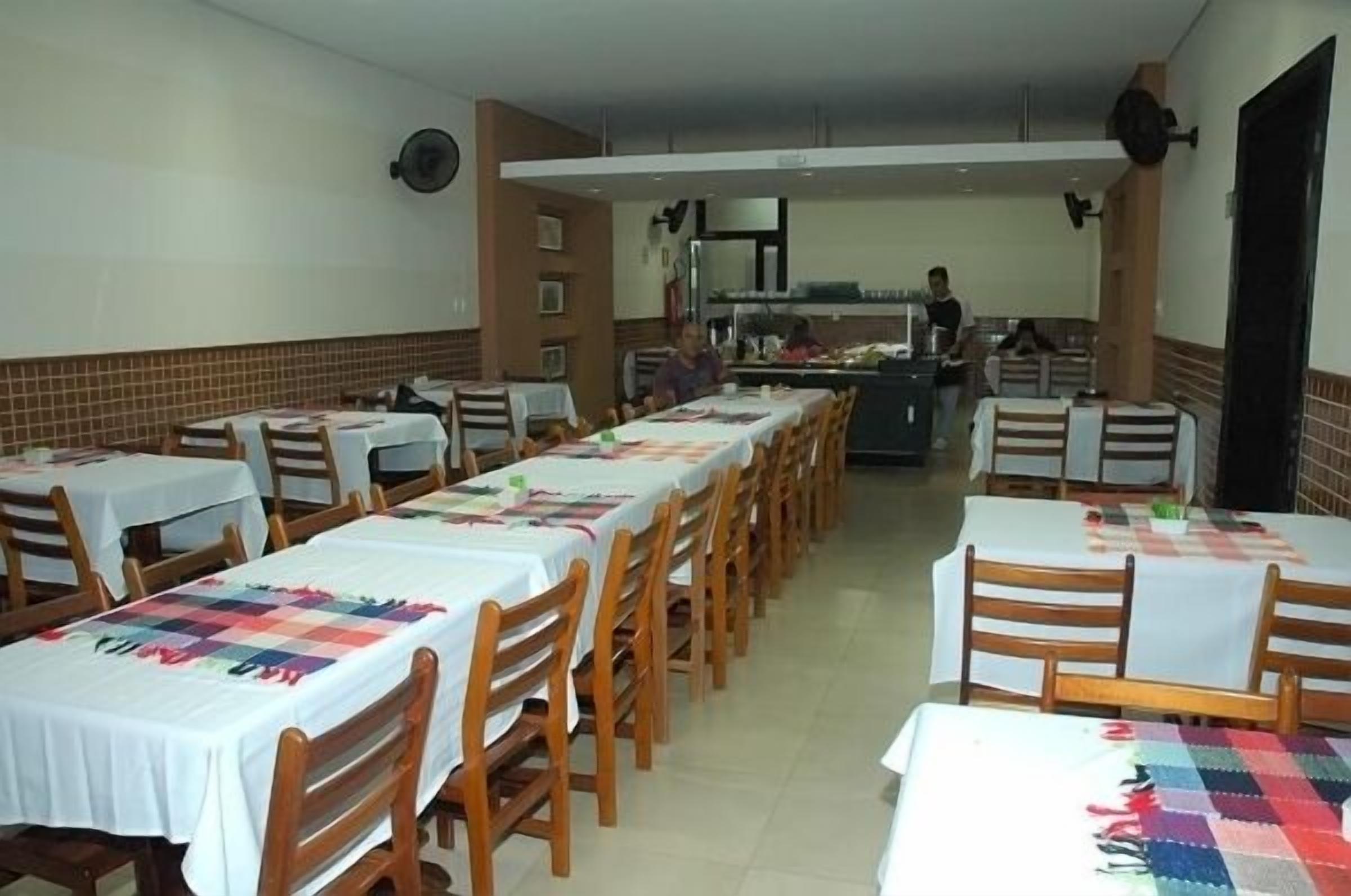 A dining area with multiple tables and chairs, a kitchen in the background, and a person working in the kitchen.