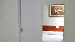 A hotel room with a white door, a bed with white bedding, a wooden headboard, and a mirror reflecting a colorful painting on the wall.