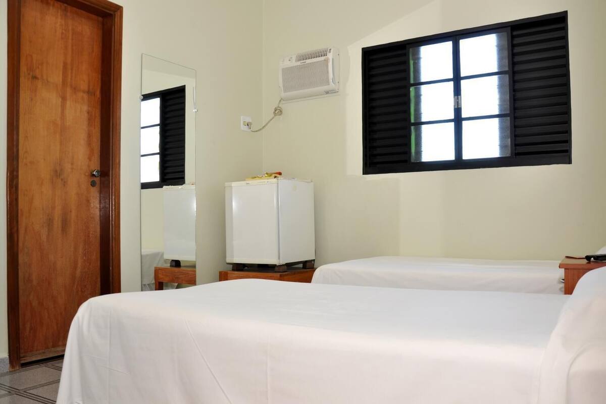 A hotel room with two beds, a wooden door, a mirror, a white air conditioner, and a window with black shutters.