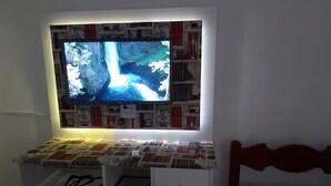 A bedroom with a TV mounted above a bookshelf, a bedside table with a lamp, and a bed with a patterned cover. - Pousada Girassol (Matinhos)