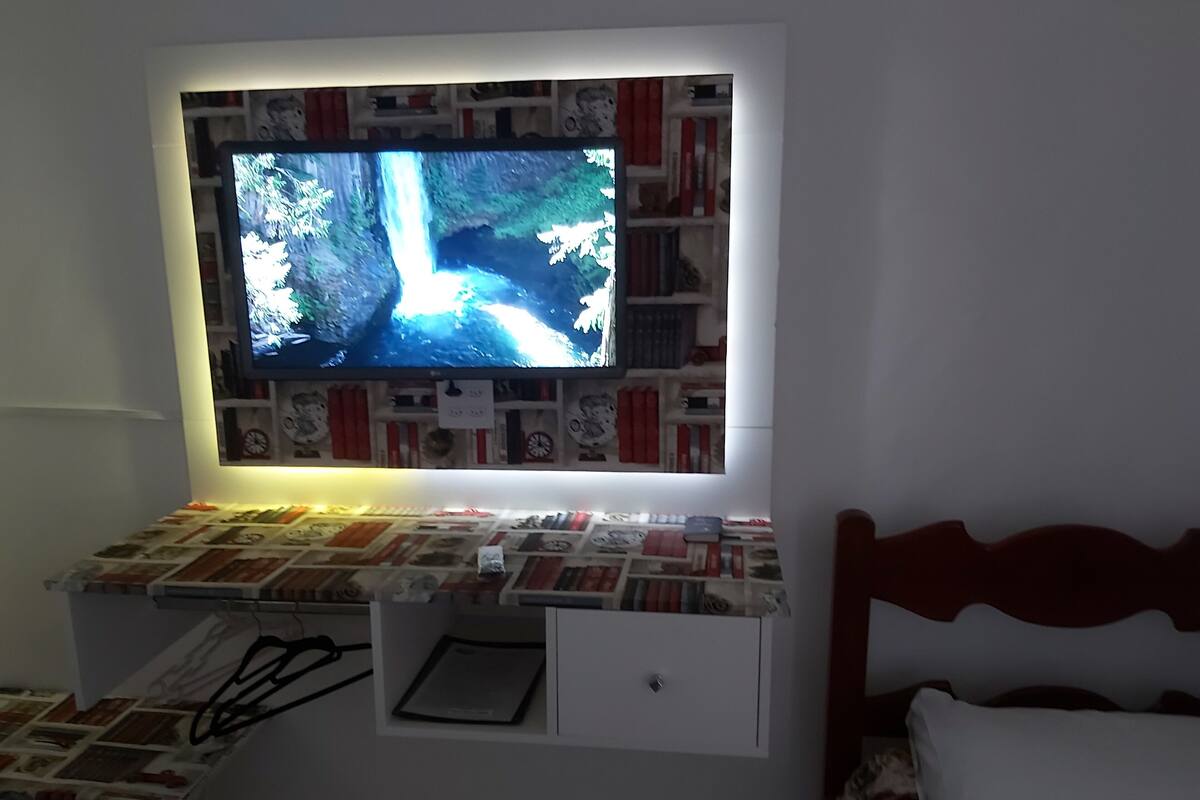 A bedroom with a TV mounted above a bookshelf, a bedside table with a lamp, and a bed with a patterned cover.