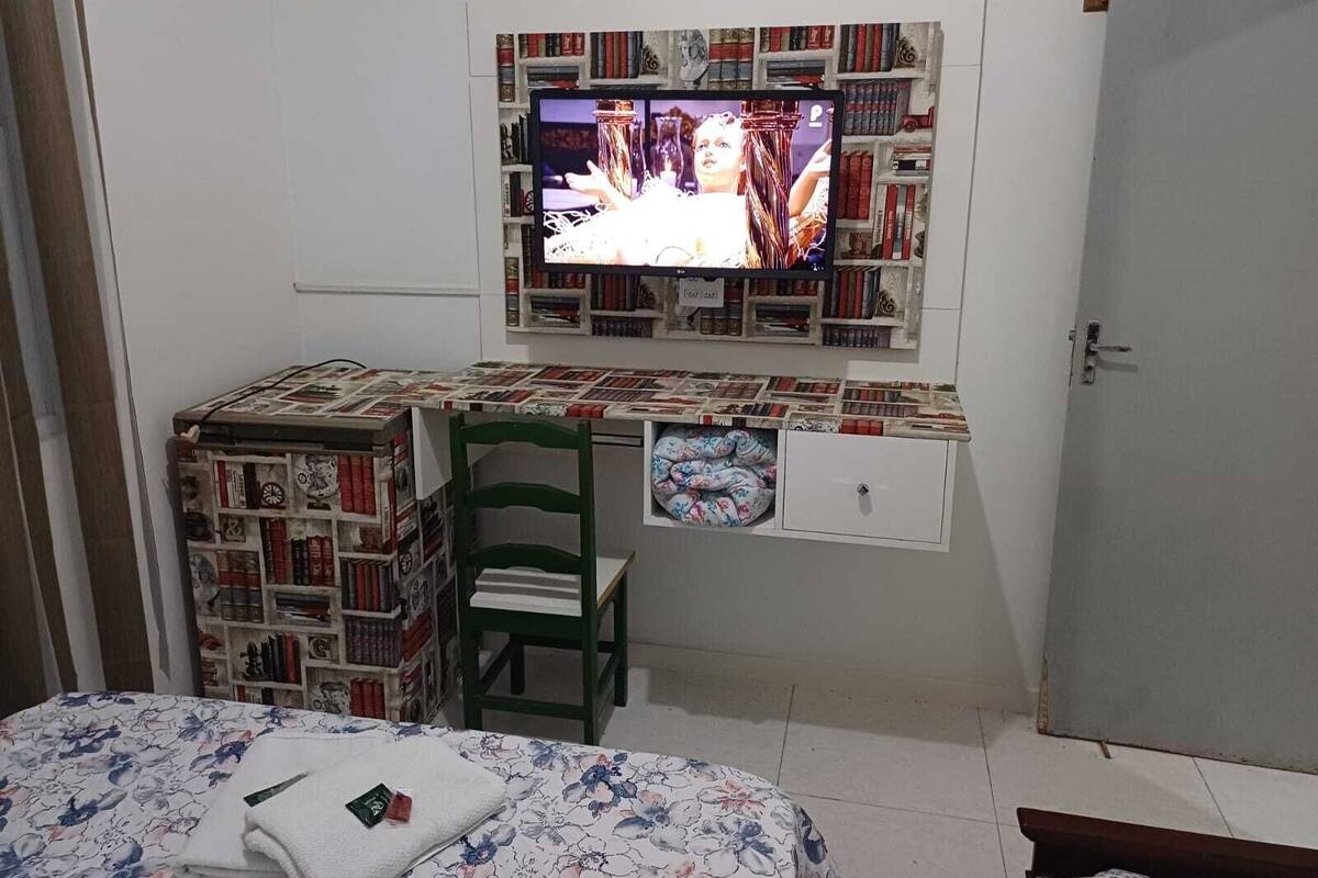 A small room with a bed, a desk with a television mounted on the wall, and a bookshelf filled with books.