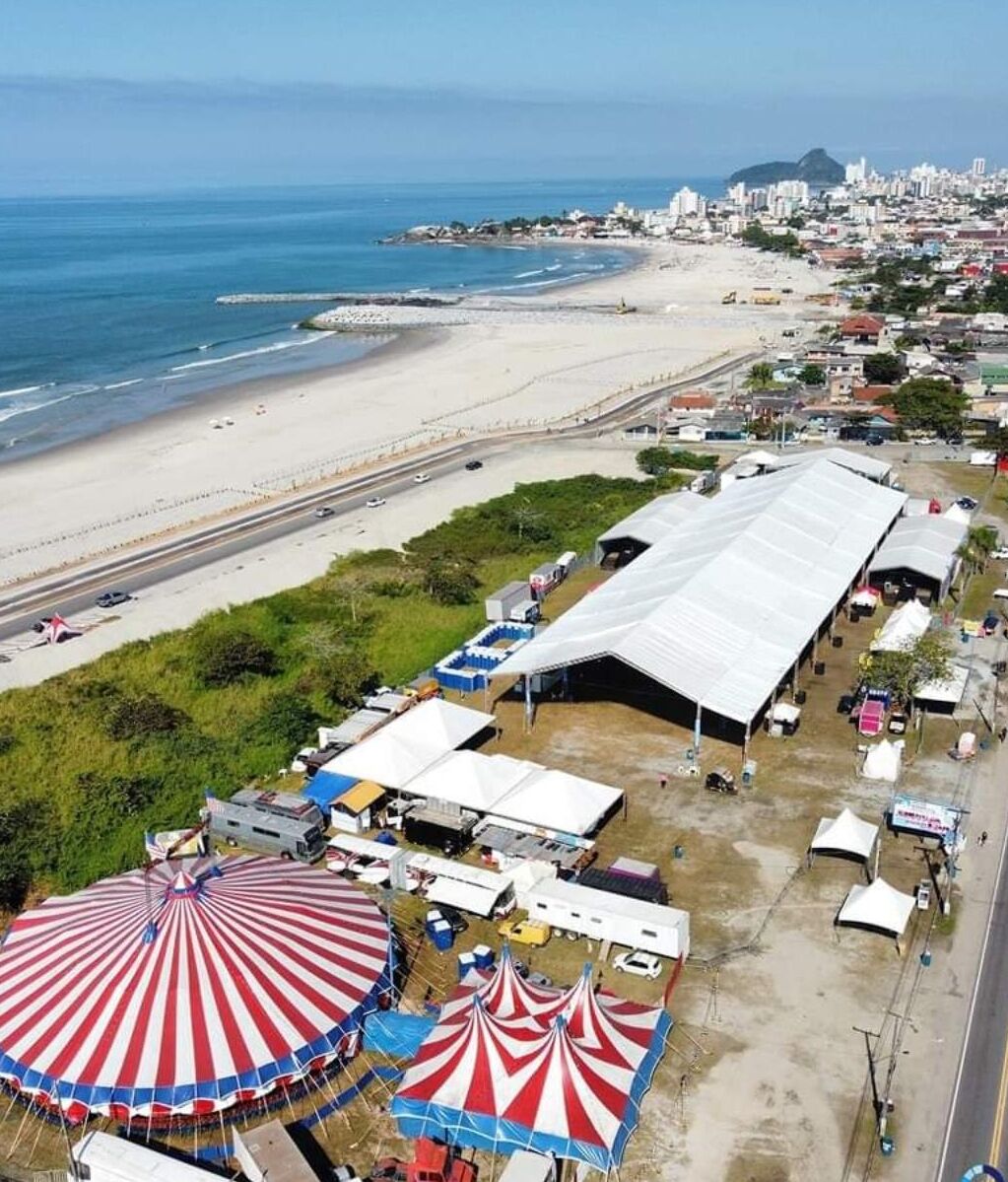 An aerial view of a beachside area with a large tented structure, a road parallel to the beach, and a cityscape in the background.