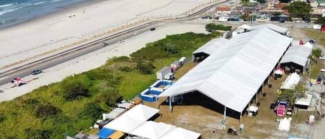 An aerial view of a beachside area with a large tented structure, a road parallel to the beach, and a cityscape in the background.