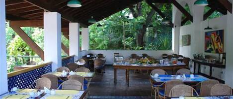 An outdoor dining area with tables and chairs, surrounded by greenery and a covered structure.