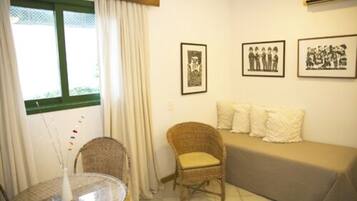 Quarto com mesa de vidro redonda, cadeira de vime e sofá com almofadas. Quadros emoldurados decoram a parede.