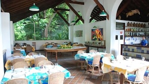 An outdoor dining area with wicker chairs and tables, a bar area with shelves of bottles, and a view of greenery through the windows. - Hotel 7 Colinas (Olinda)