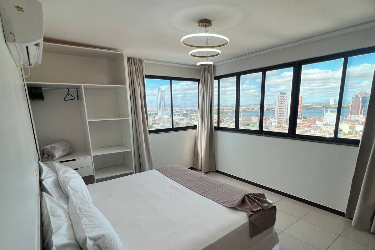 Ein modernes Hotelzimmer mit einem großen Bett, einem Kleiderschrank und Blick auf die Stadt durch große Fenster.