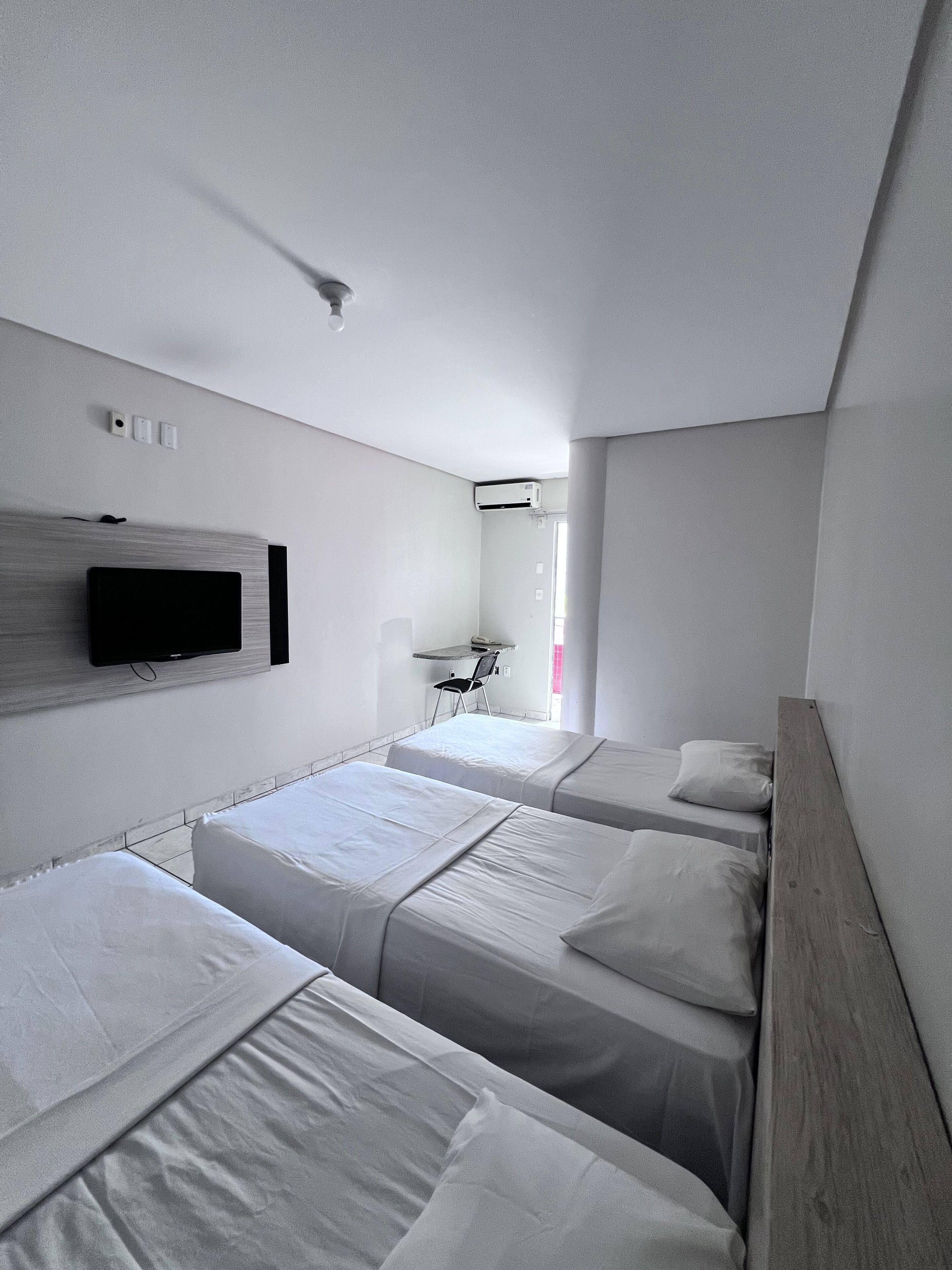 Habitación de hotel con tres camas individuales, un televisor de pantalla plana montado en la pared, un escritorio pequeño con una silla y una ventana que proporciona luz natural.
