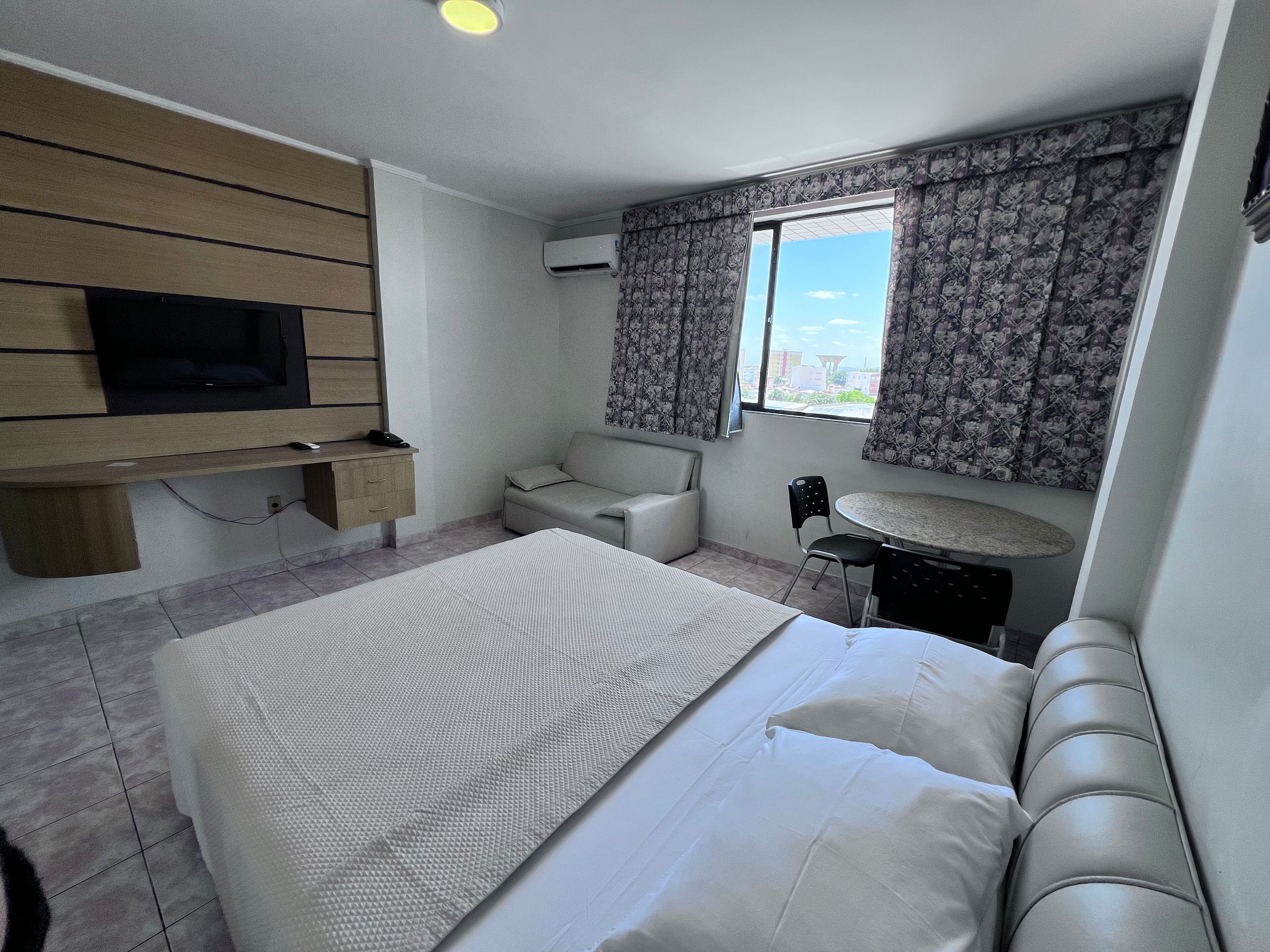 A hotel room with a bed, a TV mounted on the wall, a small table with chairs, and a window with curtains.