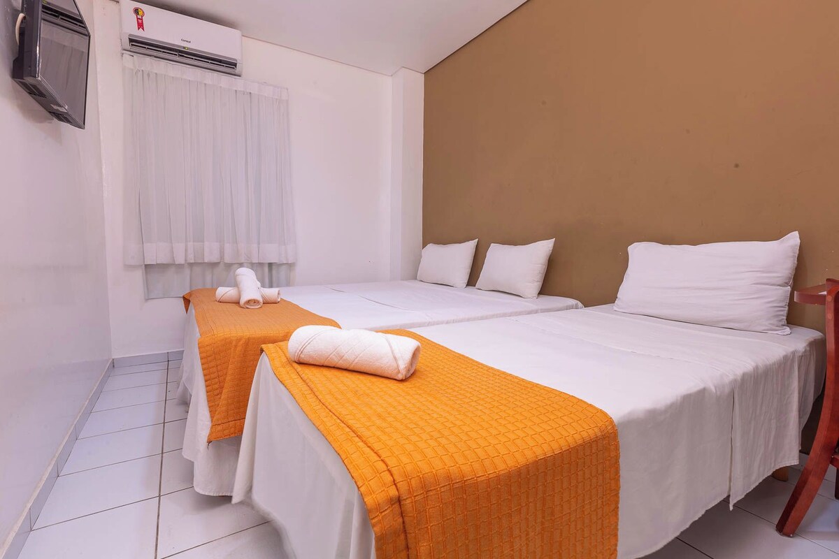 Una habitación de hotel con dos camas, ropa de cama blanca, colchas anaranjadas, una ventana con cortinas blancas y un equipo de aire acondicionado en la pared.