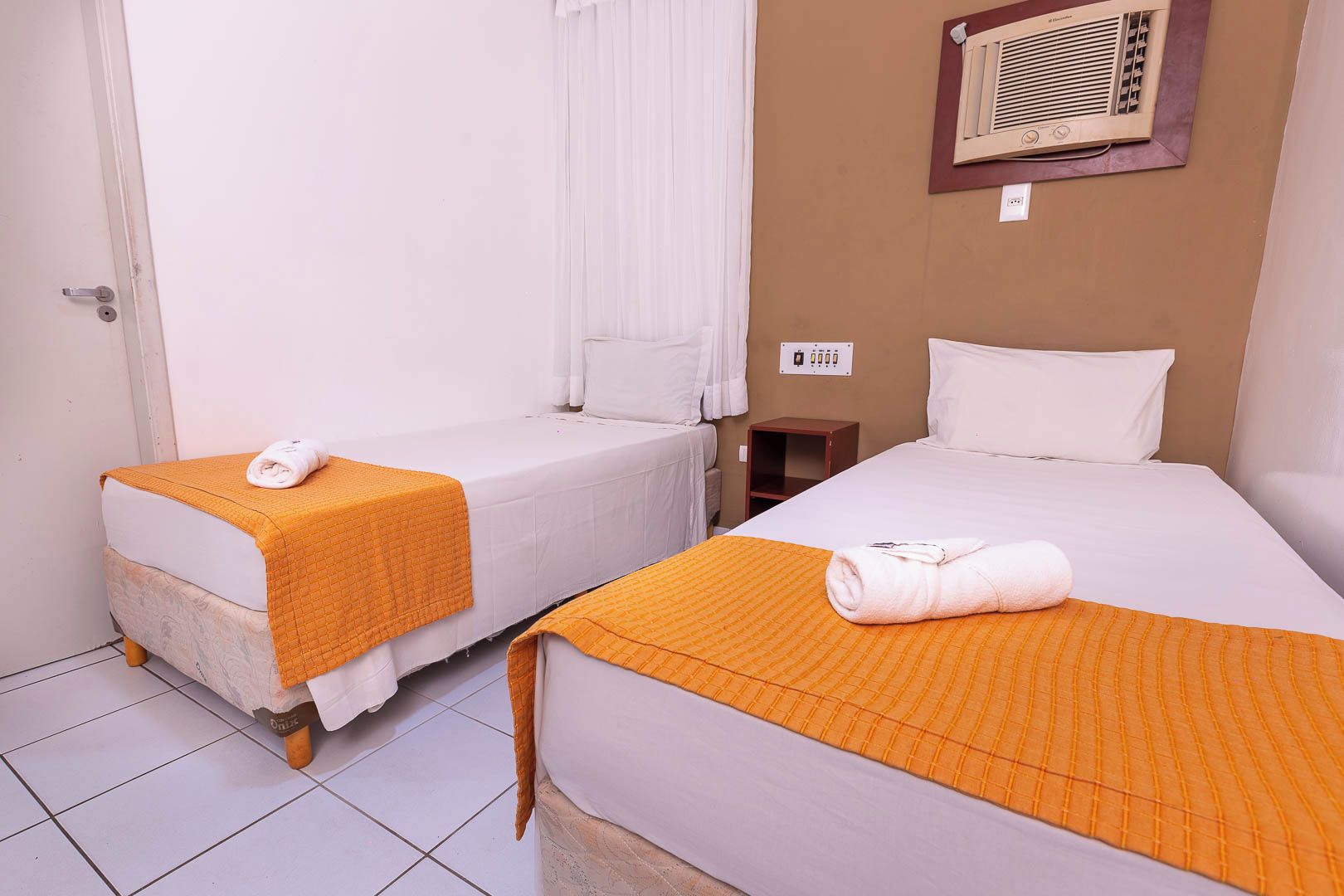 Quarto de hotel com duas camas de solteiro, cada uma com um cobertor laranja e uma toalha enrolada. Há uma pequena mesa de cabeceira de madeira e um ar-condicionado fixado na parede.