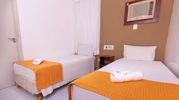 Quarto de hotel com duas camas de solteiro, cada uma com um cobertor laranja e uma toalha enrolada. Há uma pequena mesa de cabeceira de madeira e um ar-condicionado fixado na parede.