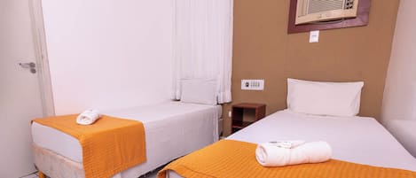 Habitación de hotel con dos camas individuales, cada una con una manta naranja y una toalla enrollada. Hay una mesita de noche de madera y un equipo de aire acondicionado instalado en la pared.