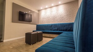 A modern living room with a sectional sofa, a flat-screen TV mounted on the wall, and a patterned wall. - Rede Andrade Navegantes (Recife)