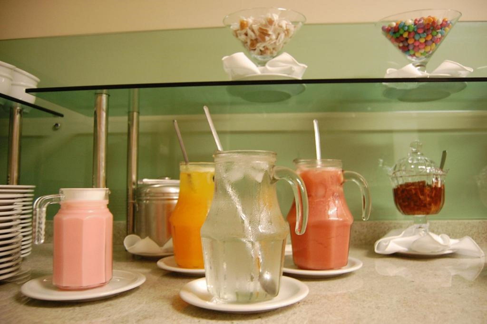 Uma estação de bebidas com diversas opções em jarras e copos, um recipiente com doces e uma pilha de pratos.