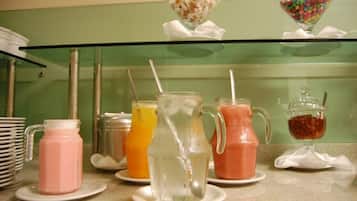 Uma estação de bebidas com diversas opções em jarras e copos, um recipiente com doces e uma pilha de pratos.