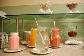Una estación de bebidas con diversas opciones en jarras y vasos, un tazón de dulces y una pila de platos.