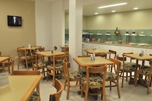 Un comedor con mesas y sillas de madera, un televisor montado en la pared y un mostrador de buffet con diversas opciones de comida.
