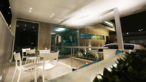 A modern outdoor dining area with white chairs and a table, surrounded by glass walls and a metal railing. - Rede Andrade Navegantes (Recife)
