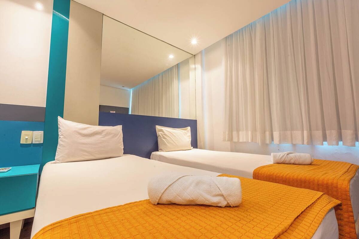 Ein Hotelzimmer mit zwei Betten, jedes mit einem weißen Kopfkissen und einer orangefarbenen Decke. An der Wand befindet sich ein Spiegel und ein Fenster mit weißen Vorhängen.