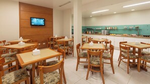 Ein Hotel-Speisebereich mit Holztischen und -stühlen, einem an der Wand befestigten Fernseher und einer Küchenzeile mit verschiedenen Speisen.