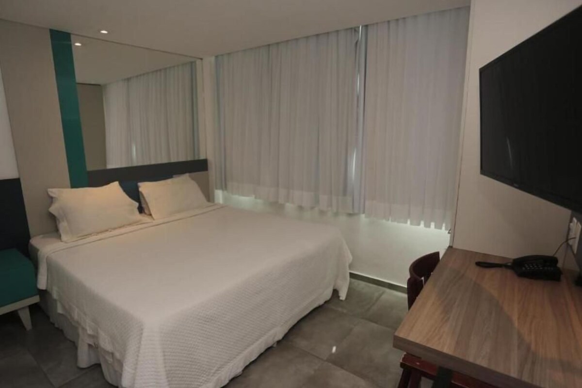 Una habitación de hotel con cama, escritorio, silla, televisor y ventana con cortinas.