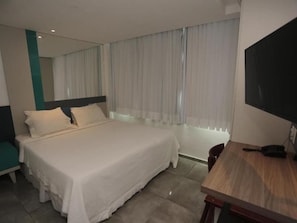 Una habitación de hotel con cama, escritorio, silla, televisor y ventana con cortinas.