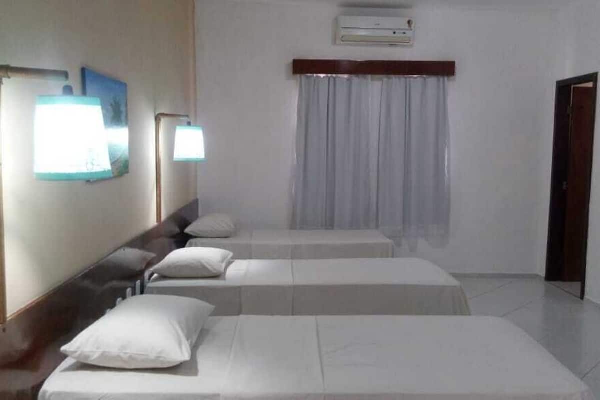 A hotel room with three single beds, a wooden headboard, a window with curtains, an air conditioning unit, and a door.