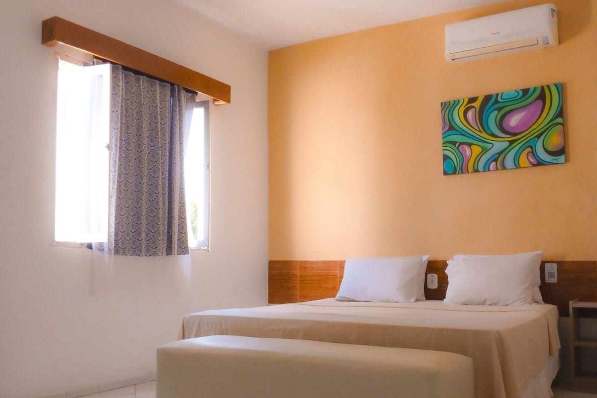 A bedroom with a bed, an ottoman, a window with curtains, an air conditioning unit, and a colorful abstract painting on the wall.