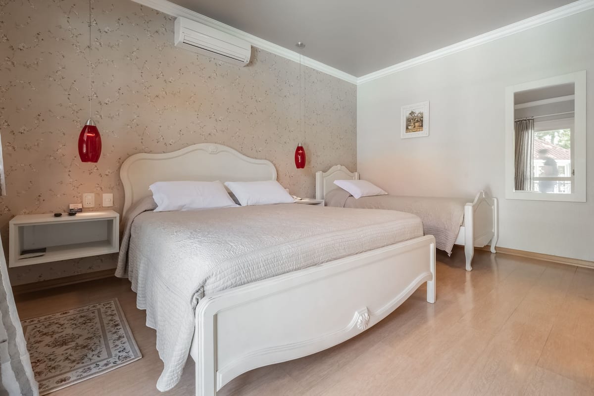 A bedroom with two beds, a nightstand, a bedside table, a wall-mounted air conditioner, a framed picture, and a window with a curtain.