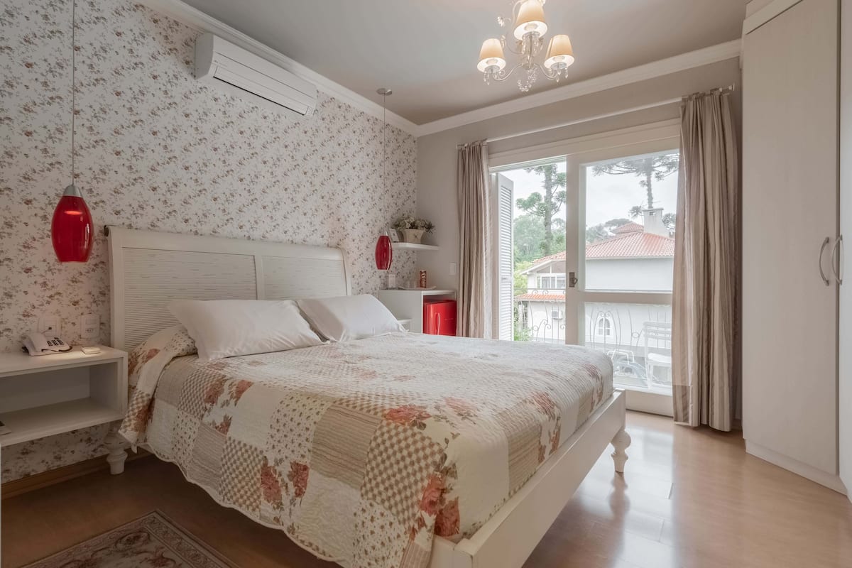 A bedroom with a bed, bedside tables, a red lamp, a window with curtains, and a view of the outside.