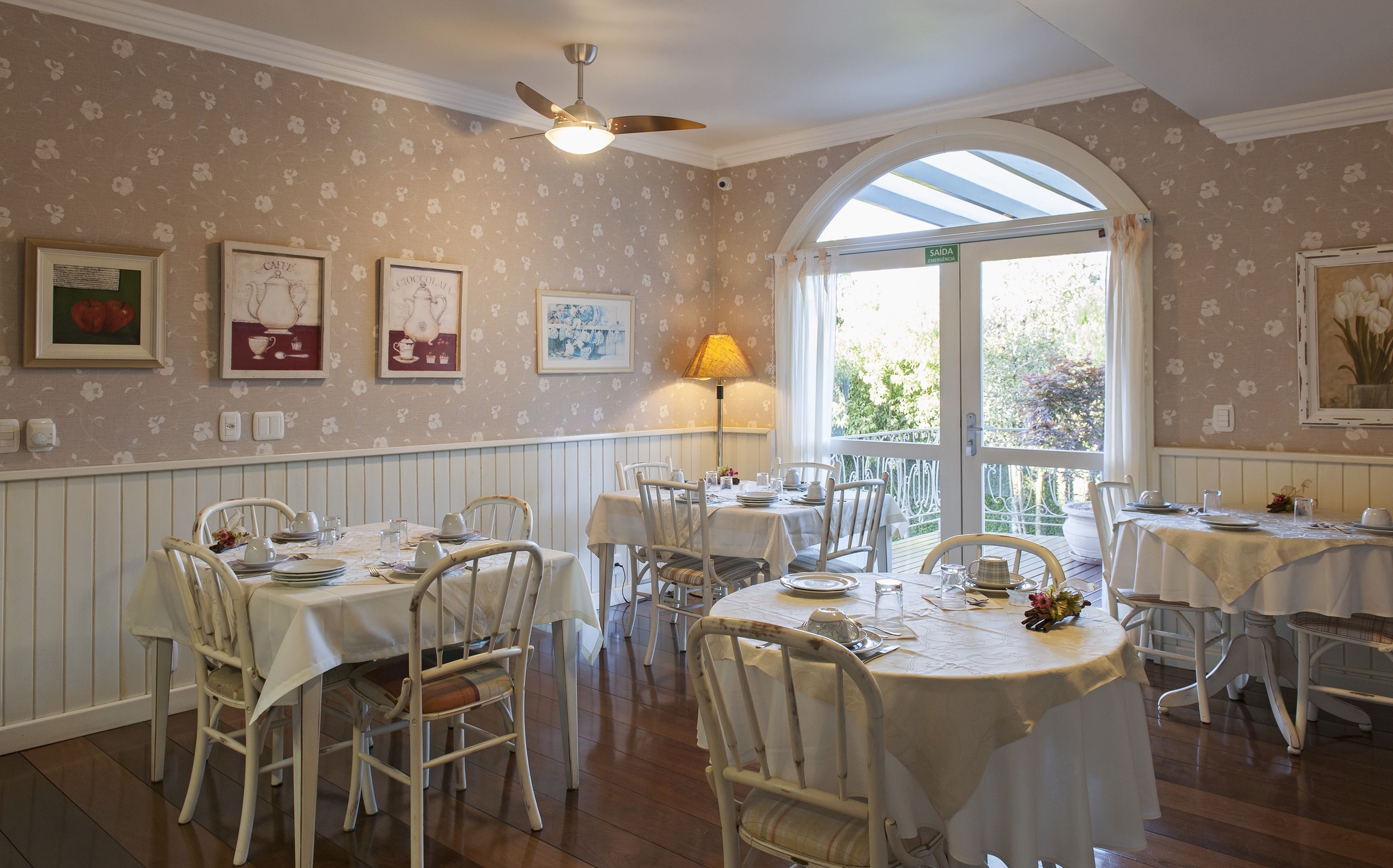 Un comedor con mesas y sillas blancas, papel tapiz floral y cuadros en las paredes.