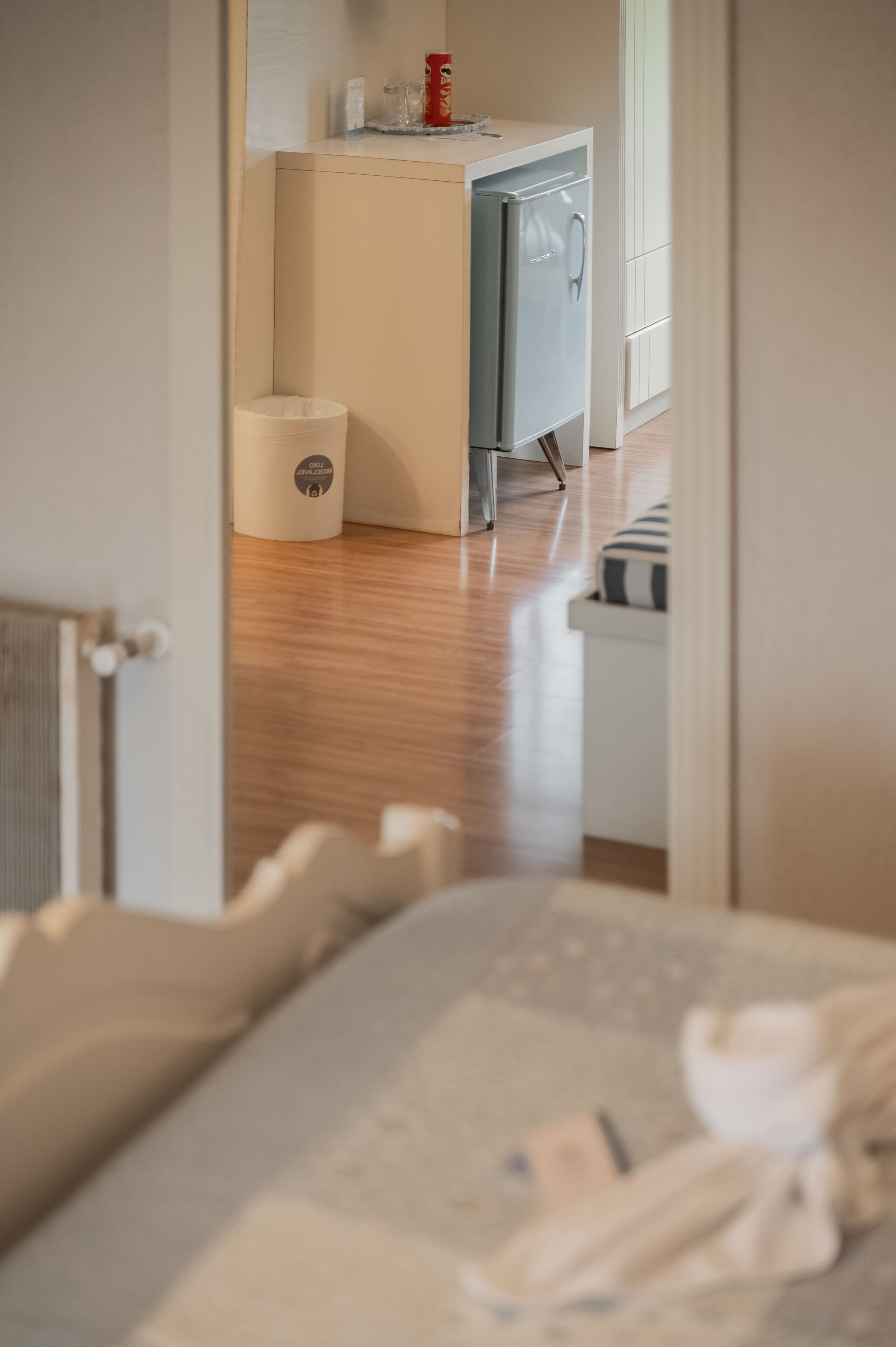 A room with a bed, a small kitchenette with a refrigerator, a trash bin, and a window.