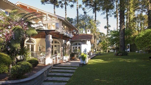 A two-story house with a stone retaining wall, a covered patio, and a well-maintained lawn. - Pousada Doce Canela (Canela)