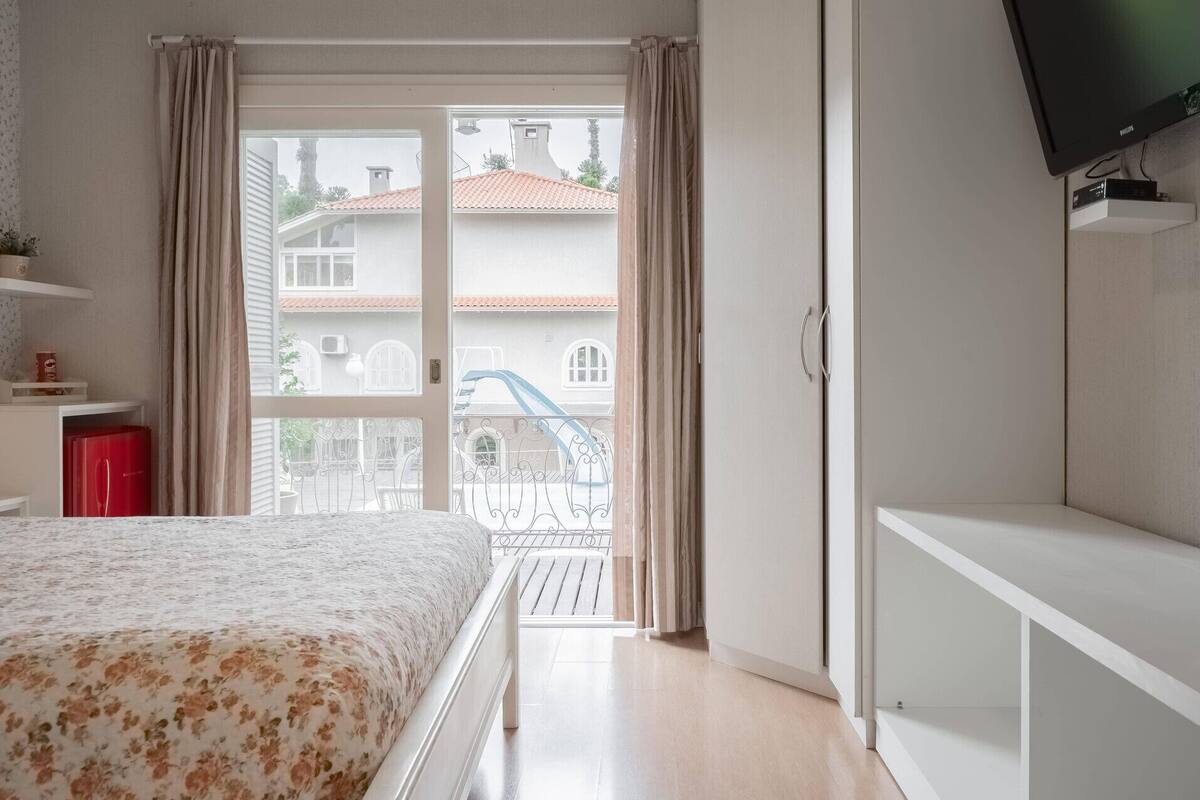 A bedroom with a bed, a TV, a balcony with a railing, and a view of buildings outside.