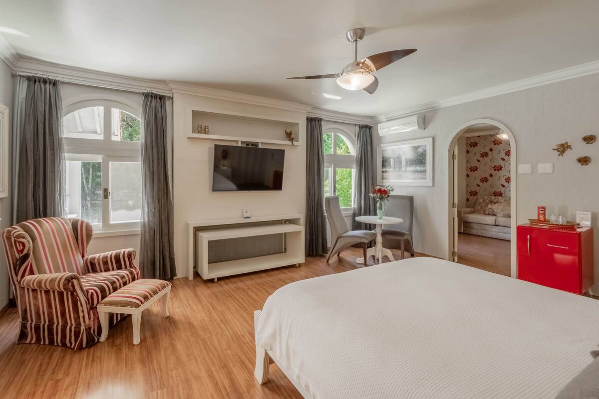 A hotel room with a bed, a TV, a chair, a small table, a ceiling fan, and a red cabinet.