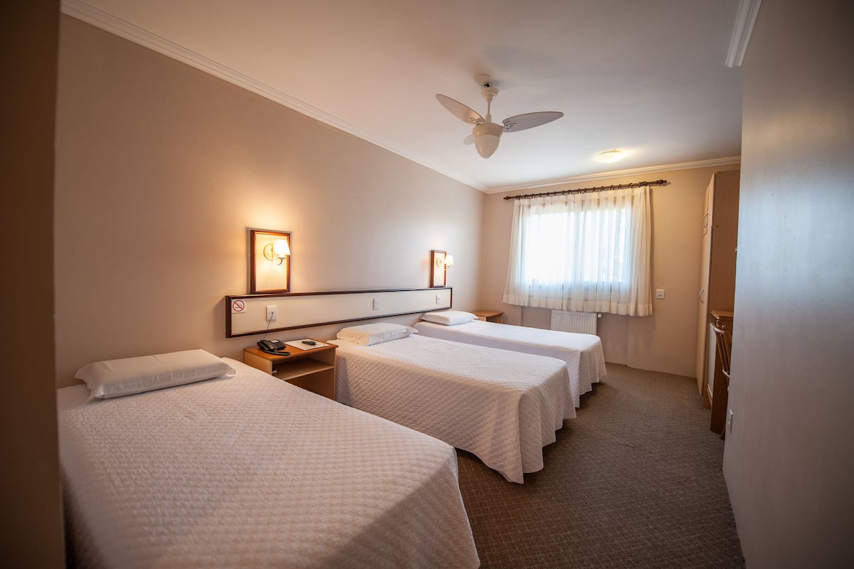 Una habitación de hotel con tres camas individuales, un ventilador de techo, lámparas de noche y una ventana con cortinas.