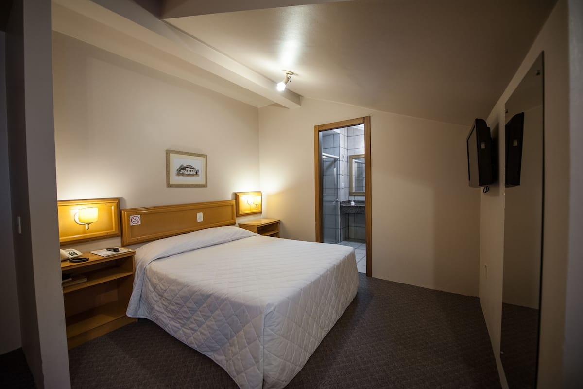 Habitación de hotel con cama, lámparas de noche, mesita de noche, un cuadro en la pared y la puerta del baño.