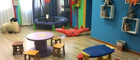 Un cuarto de juegos para niños con un trampolín, pequeñas mesas, sillas y estantes llenos de peluches.