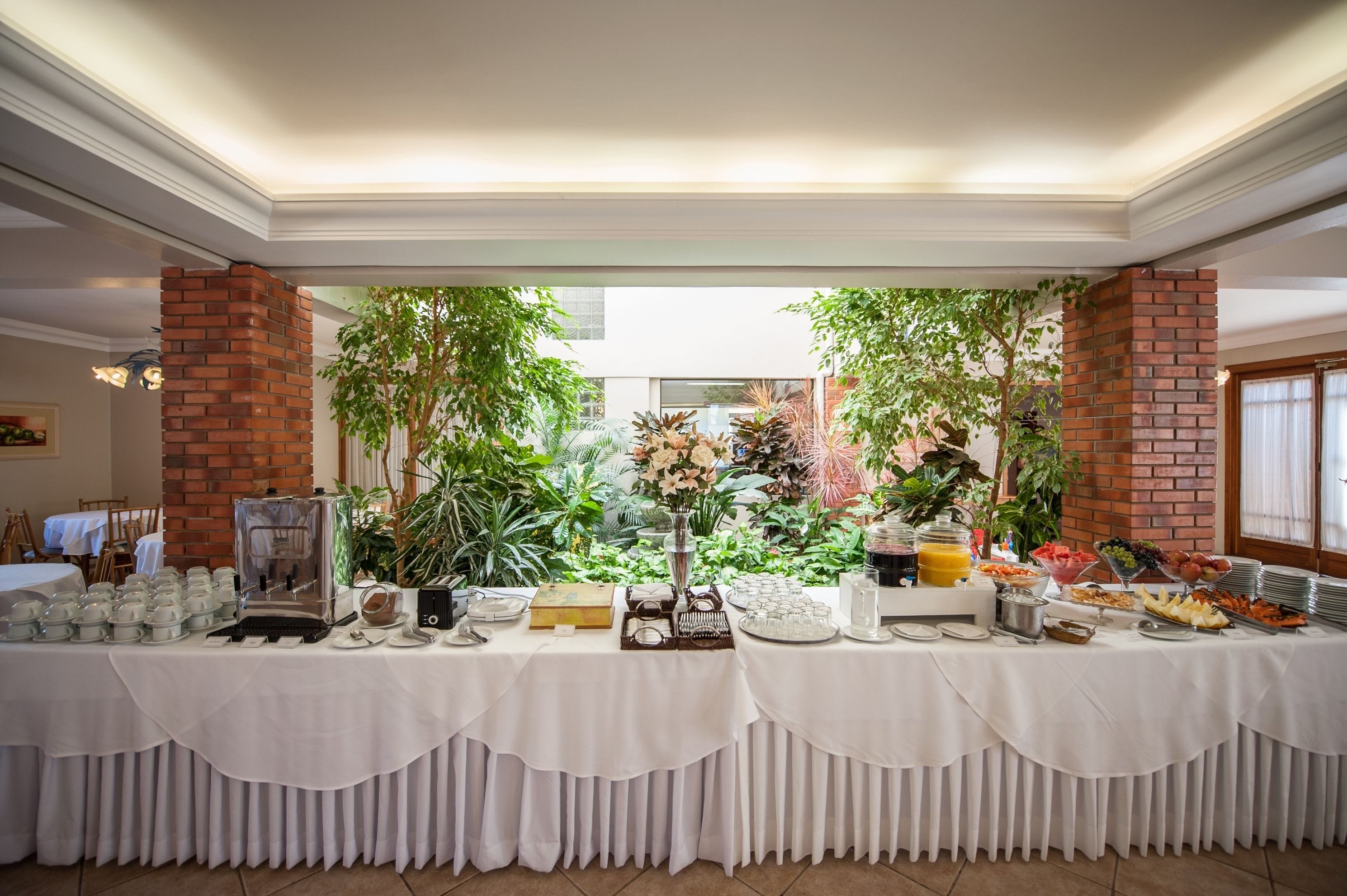 Une table de buffet proposant divers plats et un coin café, le tout agrémenté de verdure et entouré de piliers en briques.