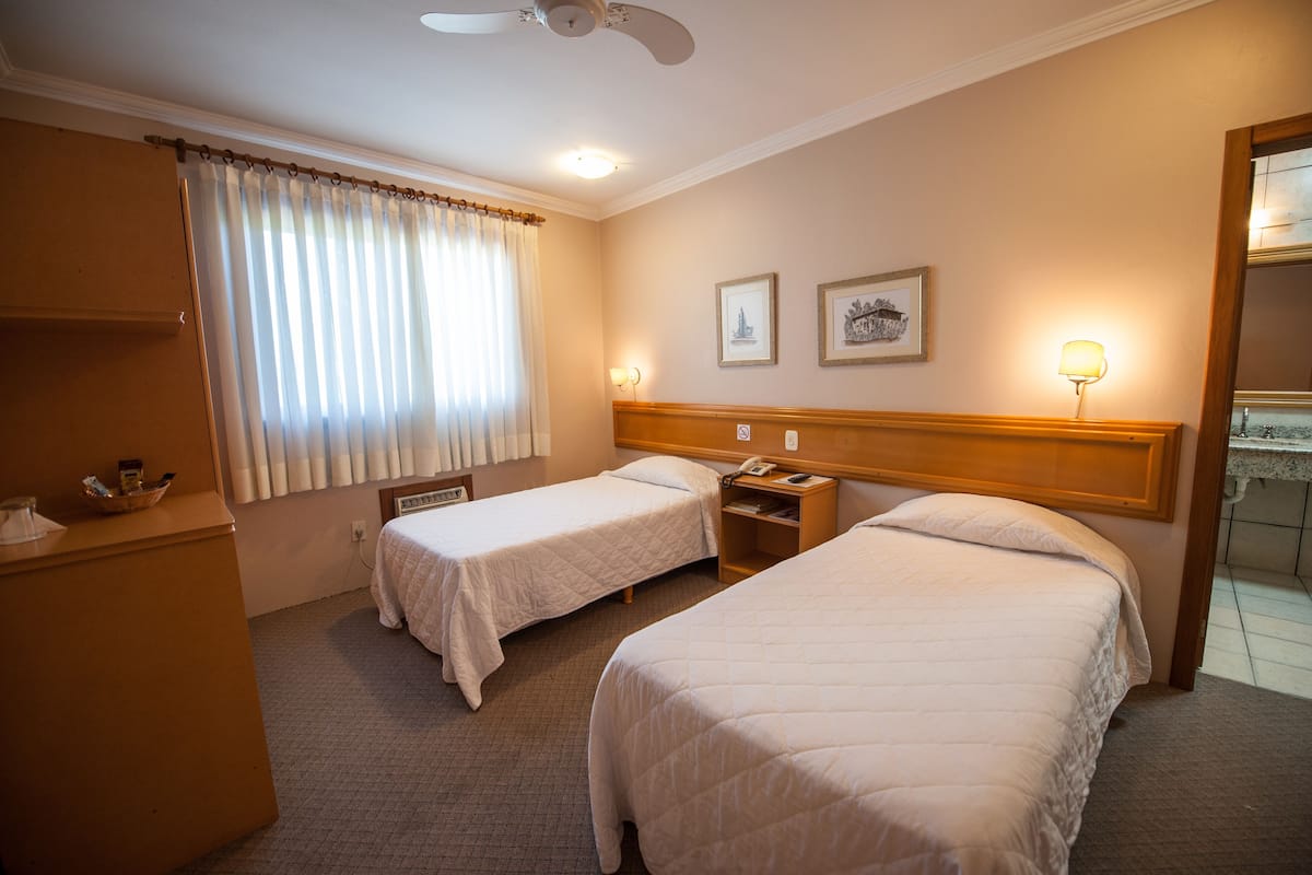 Una habitación de hotel con dos camas individuales, un cabecero de madera, una mesita de noche con lámpara, una ventana con cortinas y un ventilador de techo.